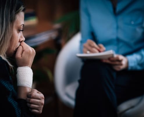 : A distressed woman with a bandaged wrist sits in a therapy session, speaking with a counselor who is taking notes.
