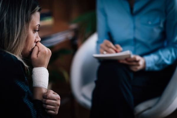 : A distressed woman with a bandaged wrist sits in a therapy session, speaking with a counselor who is taking notes.