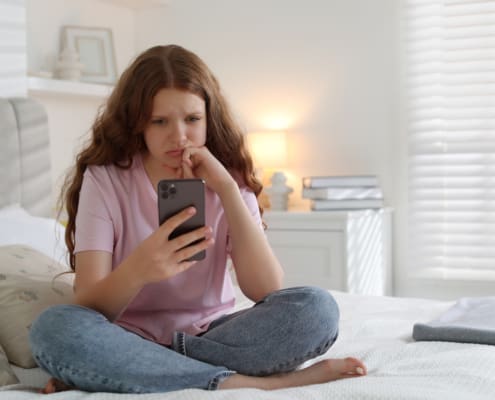 Emotional Teenage Girl Using Smartphone On Bed At Home, Space For Text