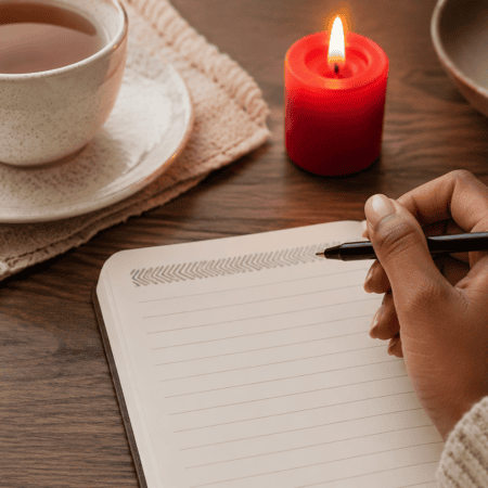 A hand writing in a notebook beside a candle and cup of tea.