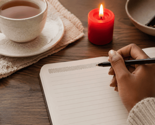 A hand writing in a notebook beside a candle and cup of tea.