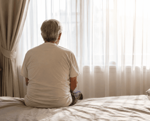 An older adult sitting on a bed facing a bright window.