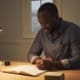 A man writing in a notebook at a wooden desk lit by a lamp, with a coffee mug beside him.
